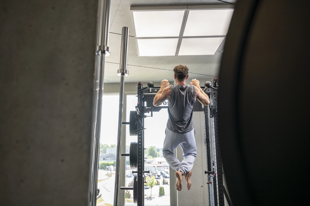 Rückansicht eines Mannes in grauer Sportkleidung, der barfuß Klimmzüge an einer Rig-Station im hellen Studio von CHEETAH Health ausführt.