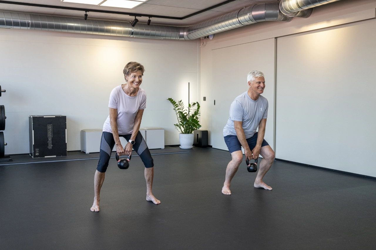 Eine Frau und ein Mann im reiferen Alter trainieren barfuß im hellen Studio von CHEETAH Health und führen lächelnd Kreuzheben (Deadlifts) mit Kettlebells aus.