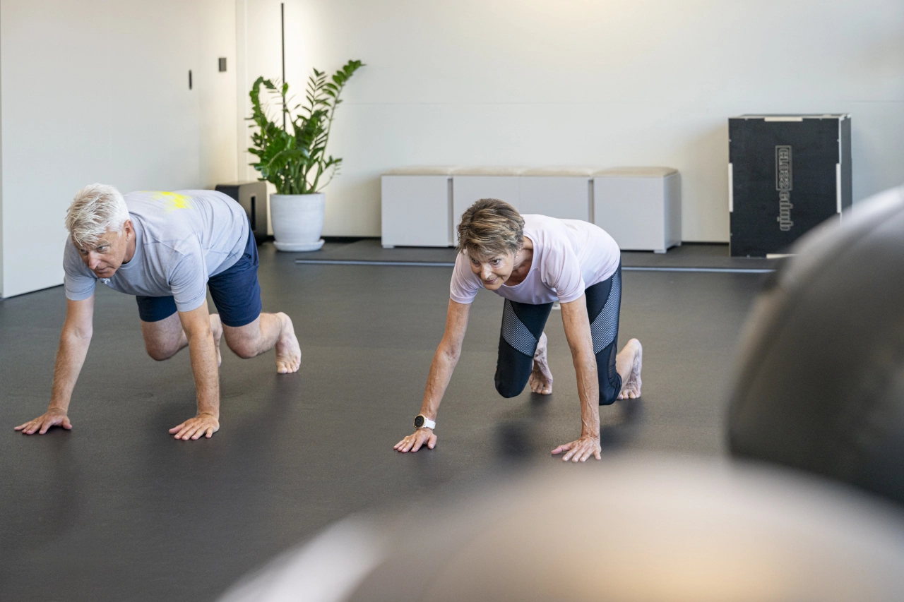 Ein älterer Mann und eine ältere Frau führen barfuß auf dem Studioboden von CHEETAH Health eine Quadrupedal-Bewegung (Bear Crawl / Bärengang) zur Stärkung der Rumpfstabilität aus.