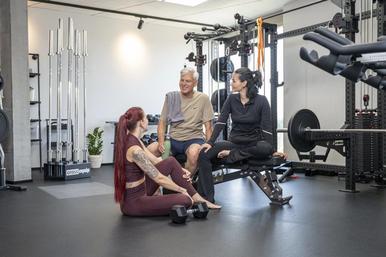 Drei Personen unterhalten sich entspannt in einer Trainingspause im hellen Studio von CHEETAH Health – eine Frau sitzt auf dem Boden, zwei weitere Personen auf einer Hantelbank.