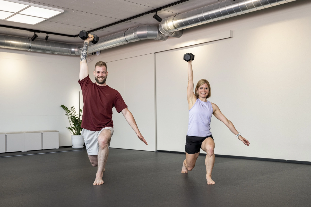 cheetah-health-funktionelles-training-kurzhantel Ein Mann und eine Frau trainieren barfuß im hellen Studio von CHEETAH Health. Sie führen synchron Ausfallschritte (Lunges) aus und stemmen dabei eine Kurzhantel einarmig über den Kopf. Der Mann trägt ein rotes T-Shirt und graue Shorts, die Frau ein fliederfarbenes Tanktop und schwarze Shorts.
