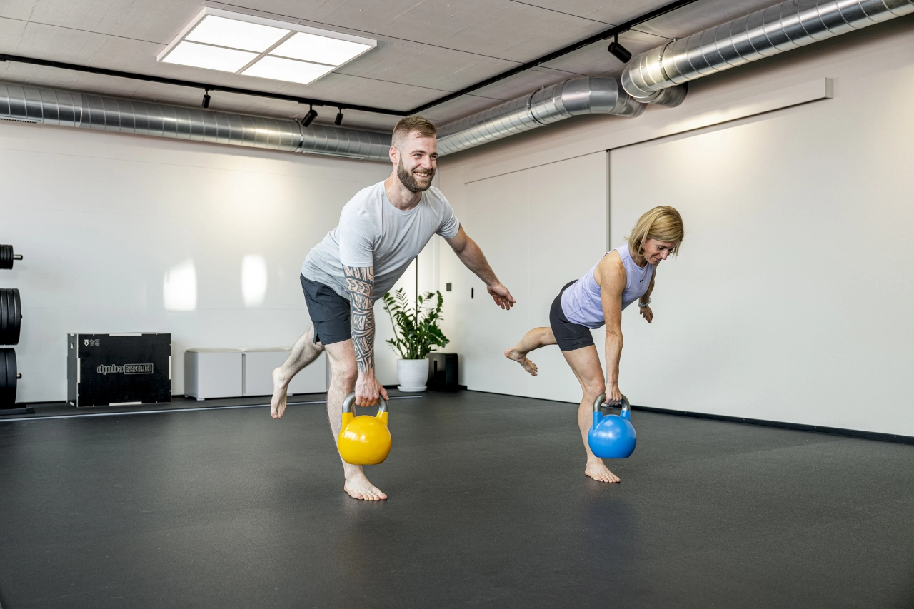 Ein Mann und eine Frau trainieren barfuß mit gelben und blauen Kettlebells (Kugelhanteln) im modernen Fitnessstudio-Bereich von CHEETAH Health.
