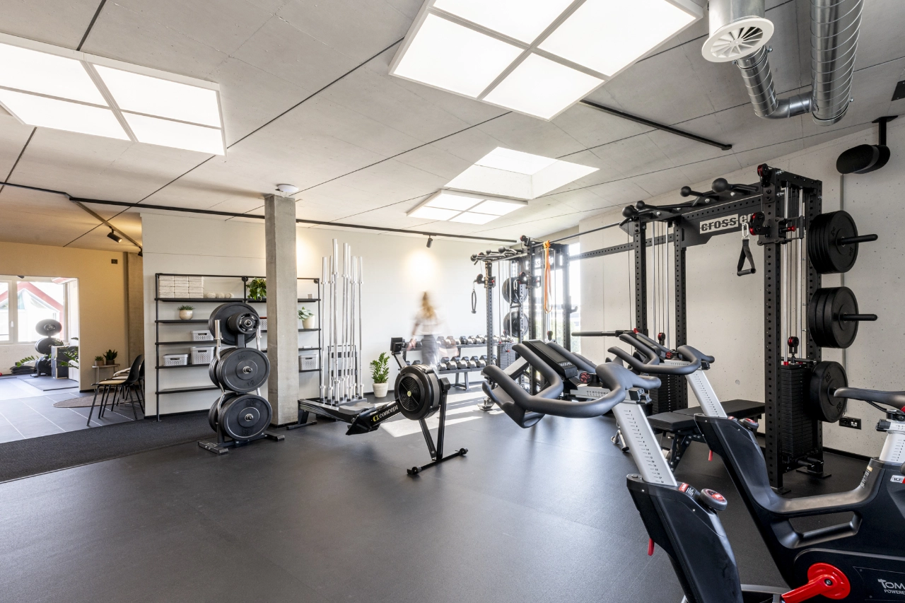 Weitwinkelaufnahme des hellen, modernen Trainingsraums von CHEETAH Health mit Ruderergometer, Ergometern, Kraftstationen und Hantelablagen.