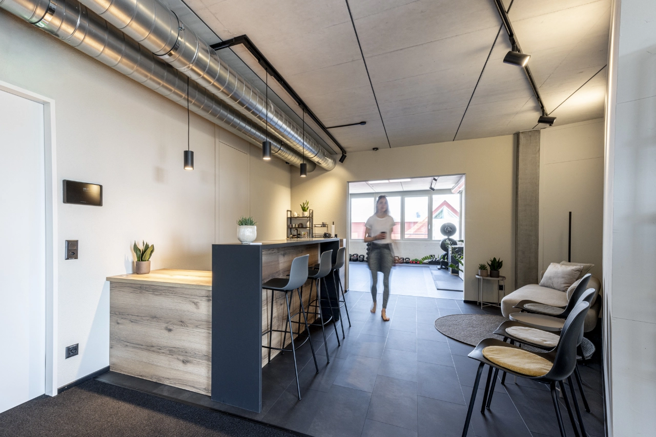 Einladender Empfangs- und Loungebereich bei CHEETAH Health mit Holztheke, Barhockern, gemütlicher Sitzecke und Blick auf den dahinterliegenden Trainingsraum.