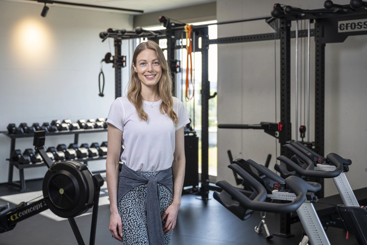 Eine lächelnde Trainerin steht im modernen Fitnessstudio von CHEETAH Health vor Trainingsgeräten wie Hantelablagen, Ruderergeometer und Kabelzug.