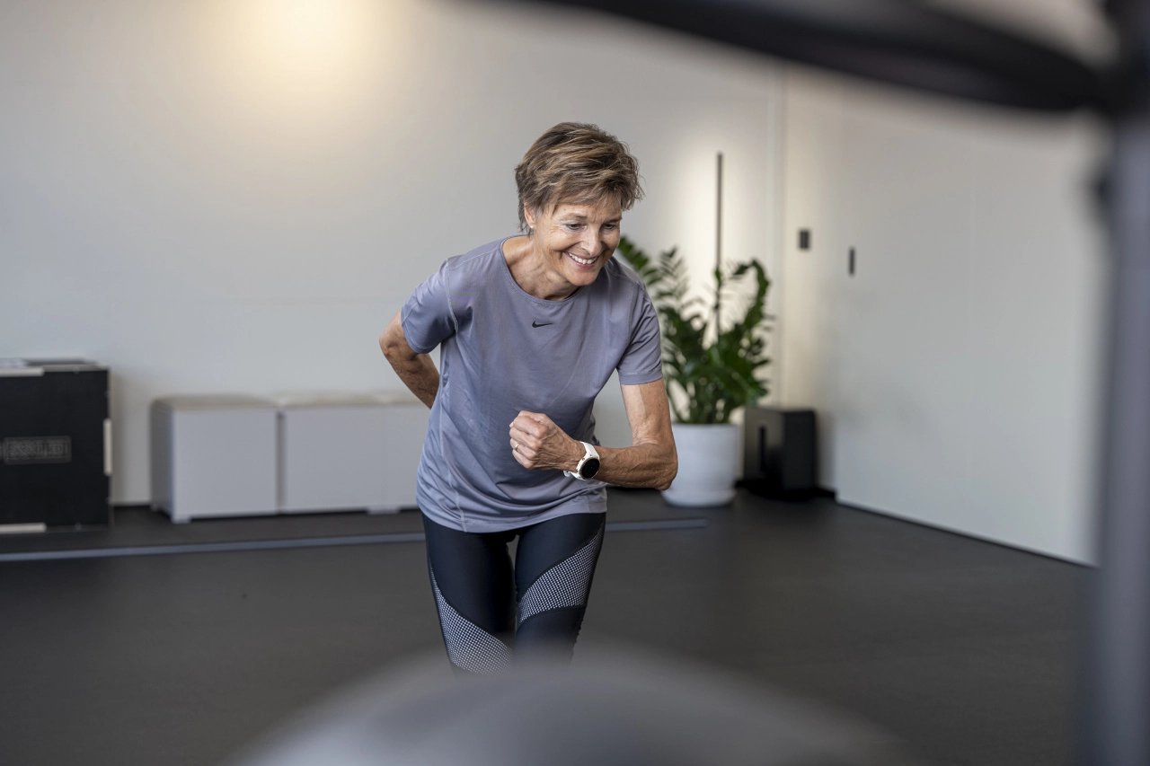 Eine lächelnde ältere Frau in grauer Sportkleidung führt dynamische Lauf- und Koordinationsübungen im hellen Studio von CHEETAH Health aus.