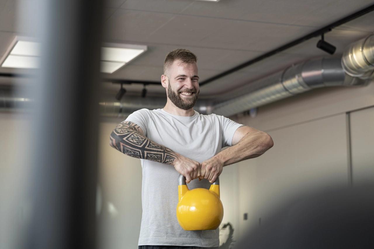 Ein lächelnder Mann mit Arm-Tattoo führt aufrecht stehend aufrechtes Rudern (Upright Row) mit einer gelben Kettlebell im Studio von CHEETAH Health aus.